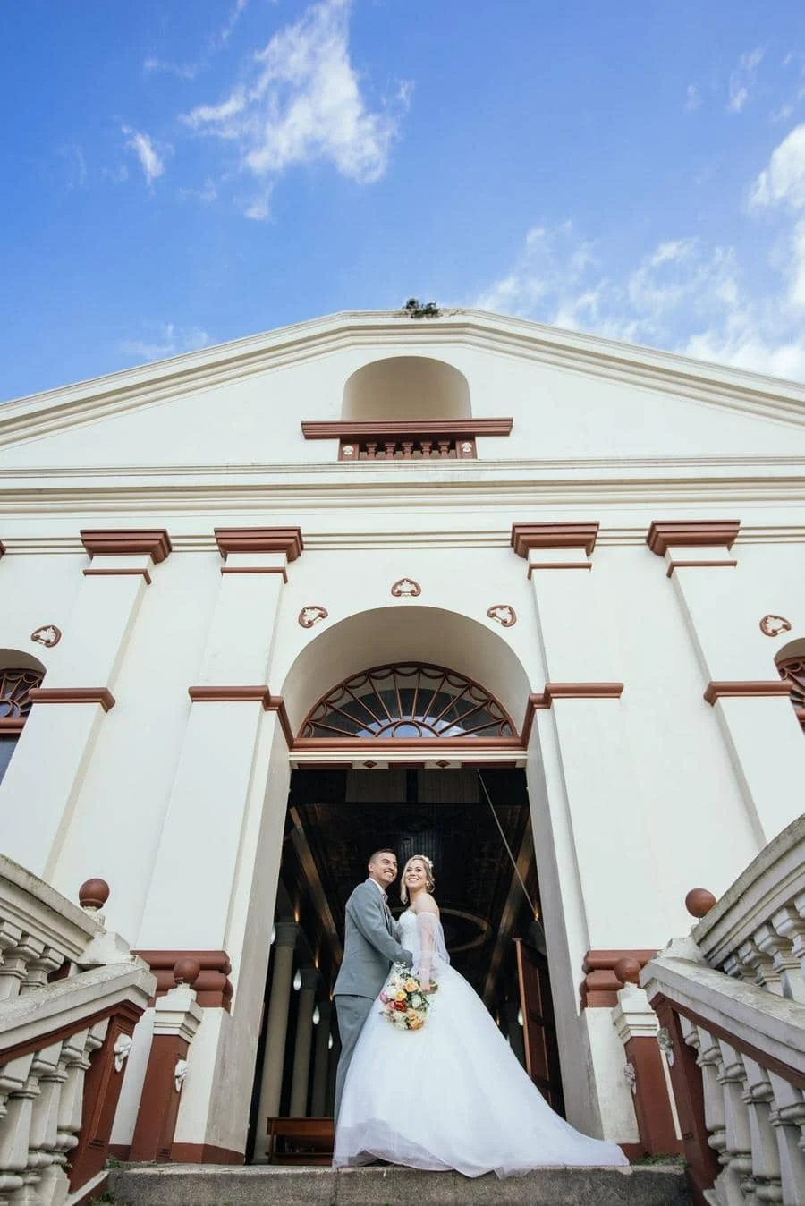 Bride and groom pose at the church entrance on stone steps, the bride holding a bouquet and wearing a white wedding gown with a long train.