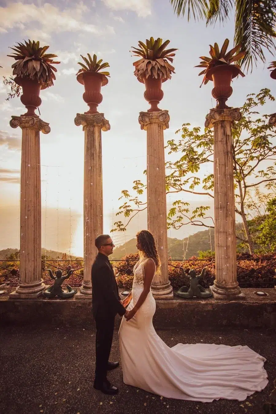 Bride in a white gown and groom in a dark suit hold hands in front of four tall stone columns at sunset.