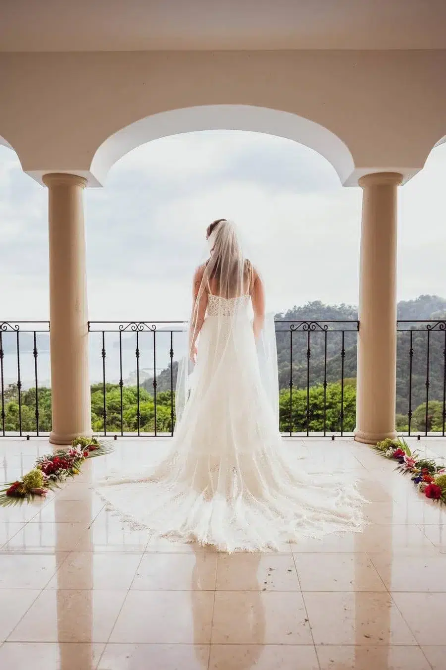 Bride in a white wedding gown with a long train and veil stands on a balcony, facing a scenic hill landscape.