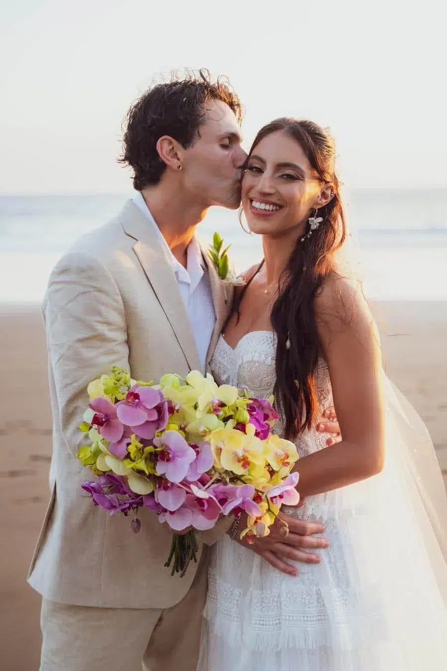Couple on the beach in wedding attire; the groom kisses the bride on the cheek as she smiles, holding a colorful orchid bouquet.