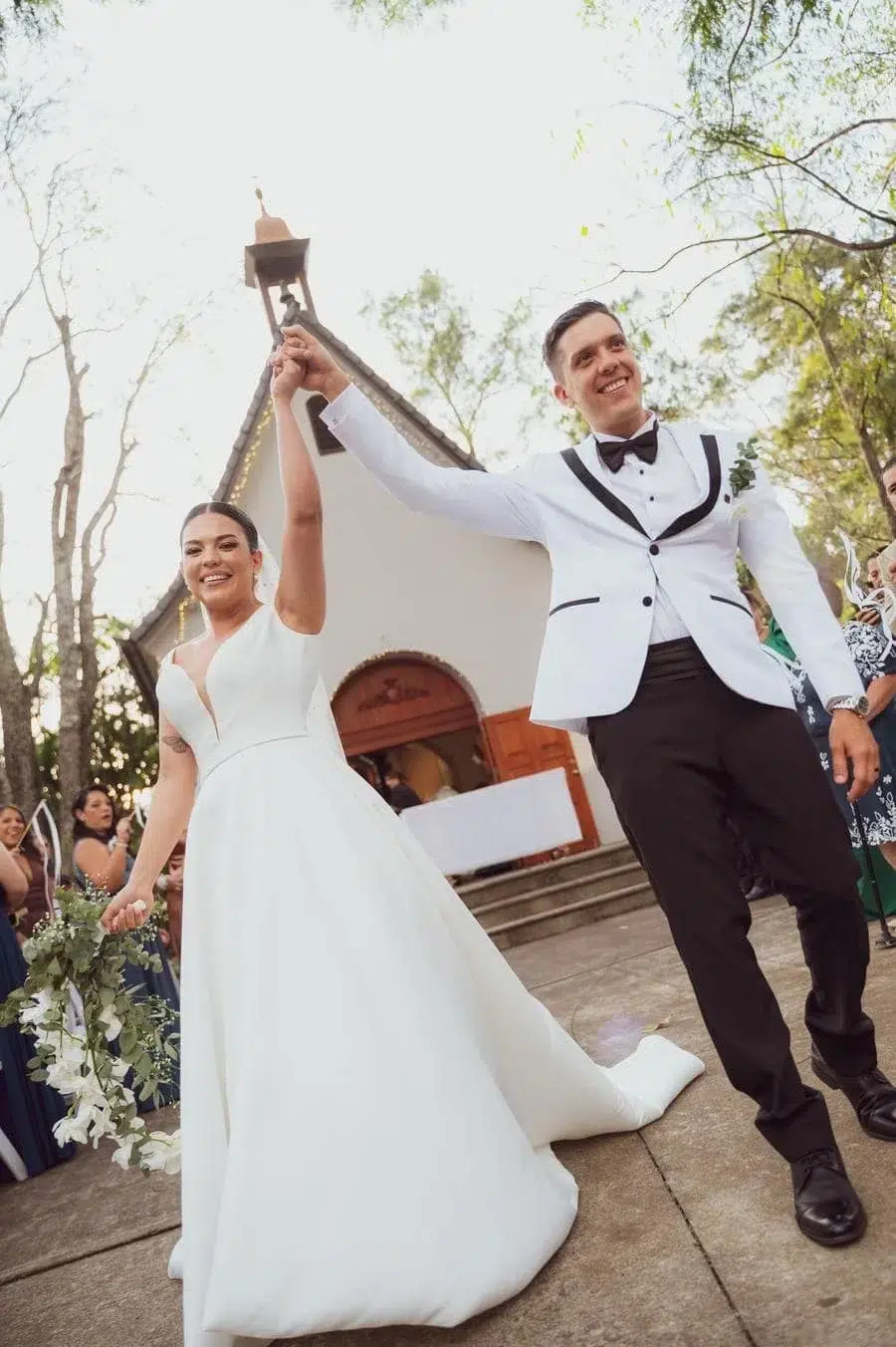 Bride and groom celebrate exiting a chapel, the bride in a white gown holding a bouquet while the groom in a white suit raises their joined hands.