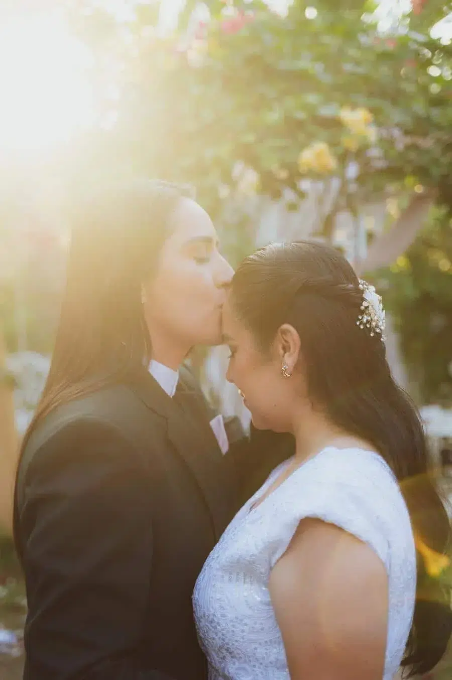 Outdoor wedding moment: one person in a dark suit kisses the forehead of another in a white lace dress as warm sunlight glows between them.