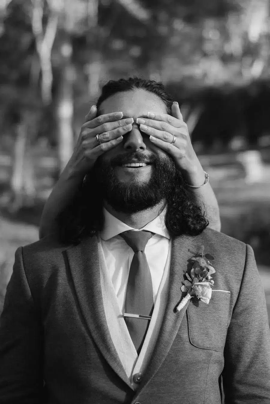 Groom in a suit with a boutonniere smiles as someone covers his eyes playfully from behind.