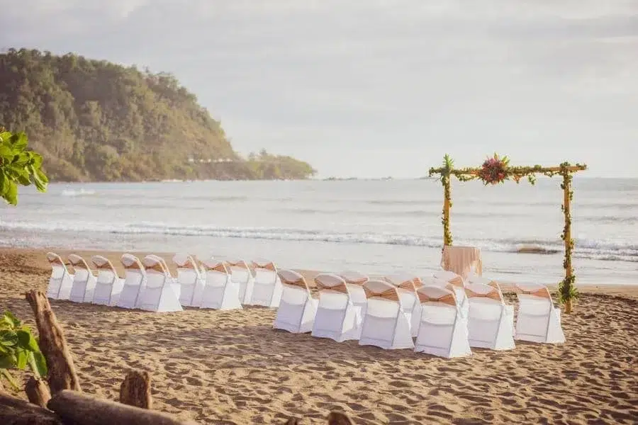 wedding ceremony photography costa rica-4 | Pacific Photography Beach wedding setup on the sand with white chairs in rows facing a flower-adorned arch by the sea.