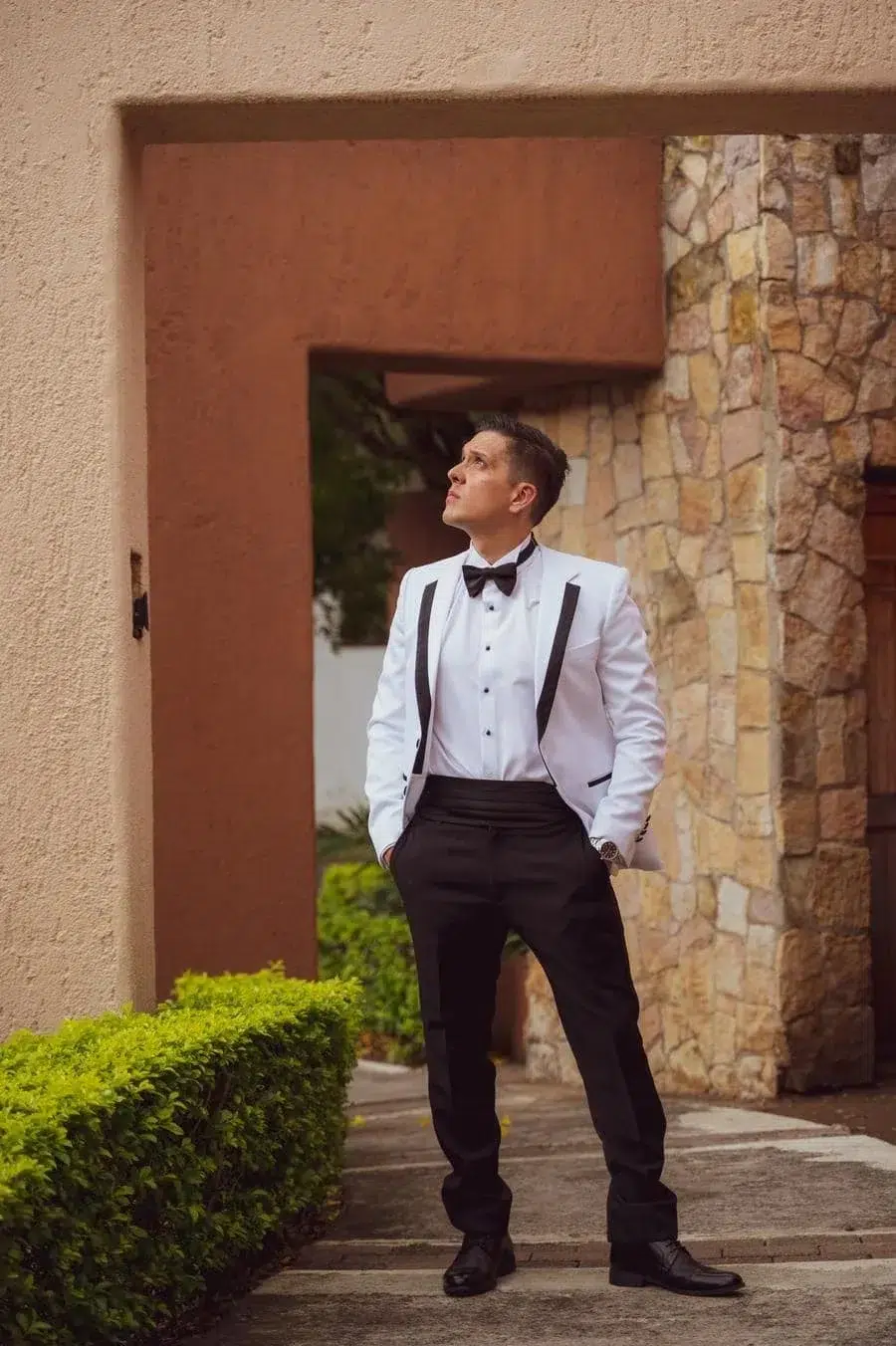 Man in a white tuxedo jacket, black bow tie, and black pants stands outdoors beside a stone wall, hands in pockets, looking up.