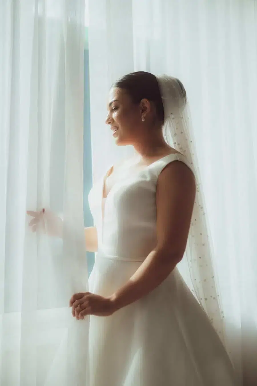 Bride in a white wedding gown and veil stands by a window, looking out.