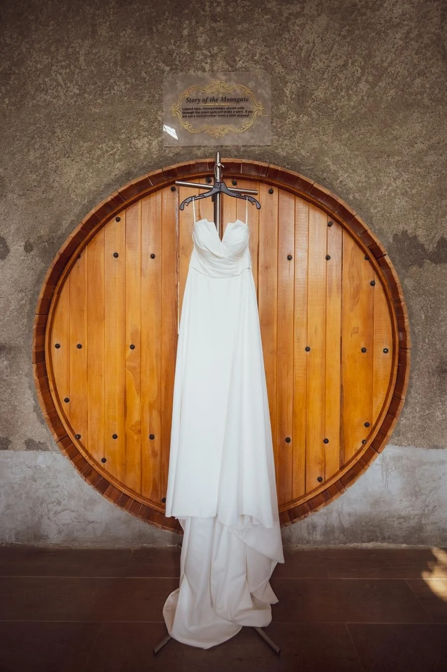 White wedding dress on a hanger in front of a large round wooden door with a sign above that reads 'Story of the Moongate'.