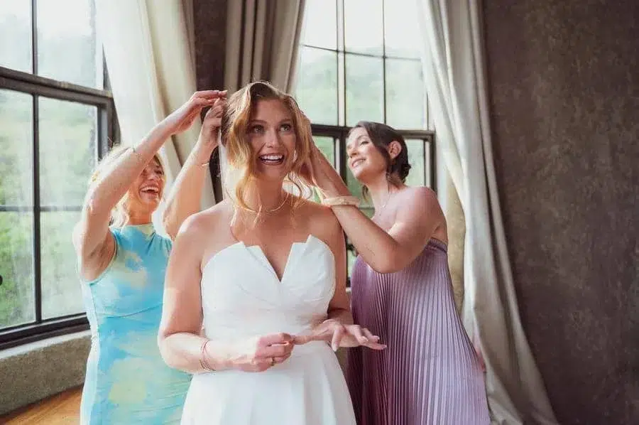 wedding prep photography costa rica-23 | Pacific Photography Bride in a white wedding dress smiles as two bridesmaids adjust her hair in a sunlit room by a large window.