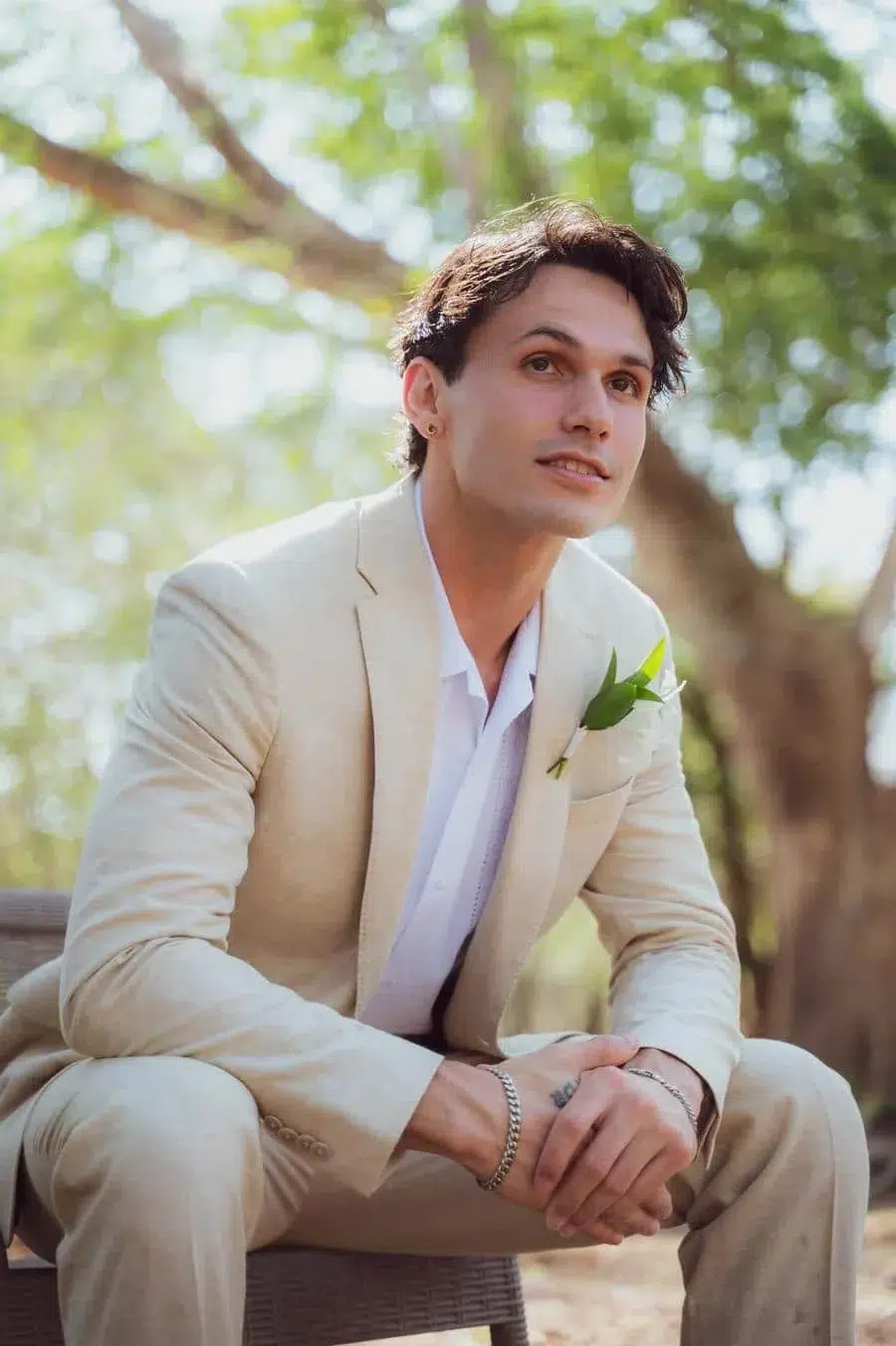 Man in a light beige suit with a white shirt and boutonniere sits on a bench outdoors, hands clasped.