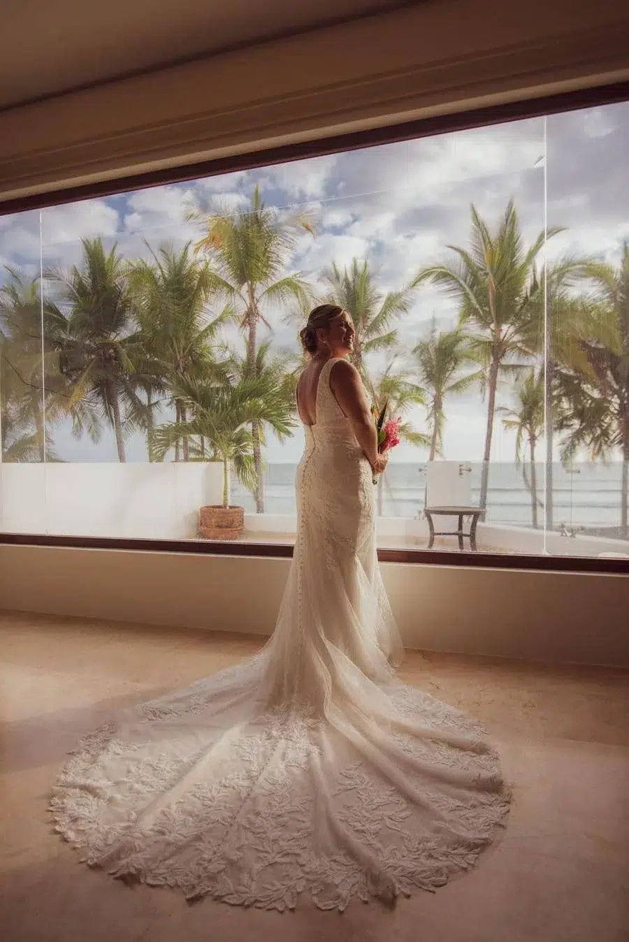 Bride in a lace wedding gown with a long train standing indoors by a large window, looking toward the ocean and palm trees outside.